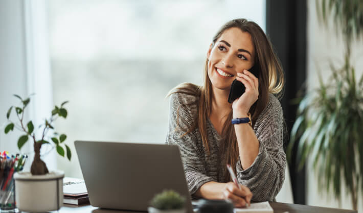 Mulher sorrindo falando ao telefone, anotando em um bloco de notas com uma caneta